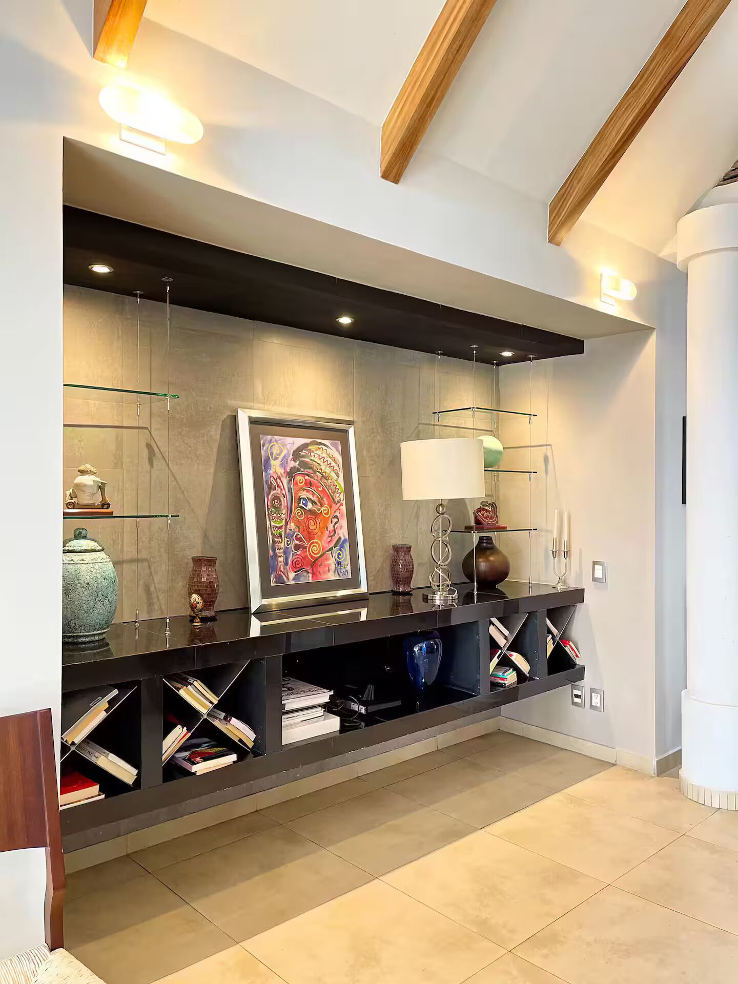 A modern interior with a built-in console table displaying books and decorative items, a framed painting, and a wall-mounted lamp.