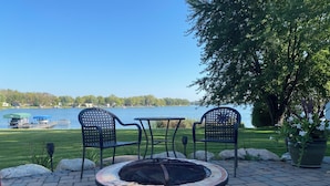 Un patio con dos sillas y una mesa, un brasero y vistas a un lago con un muelle y barcos.