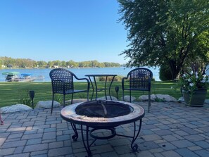 Eine Terrasse mit zwei Stühlen, einem Tisch, einer Feuerstelle und Blick auf einen See mit einem Steg und Booten.