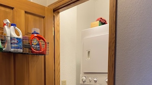Un lavadero con lavadora, un estante con productos de limpieza y una puerta de madera.