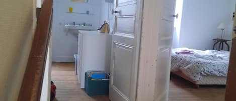 Una habitación con suelo de madera, cama con ropa de cama estampada, una puerta blanca y una mesita de noche con lámpara.