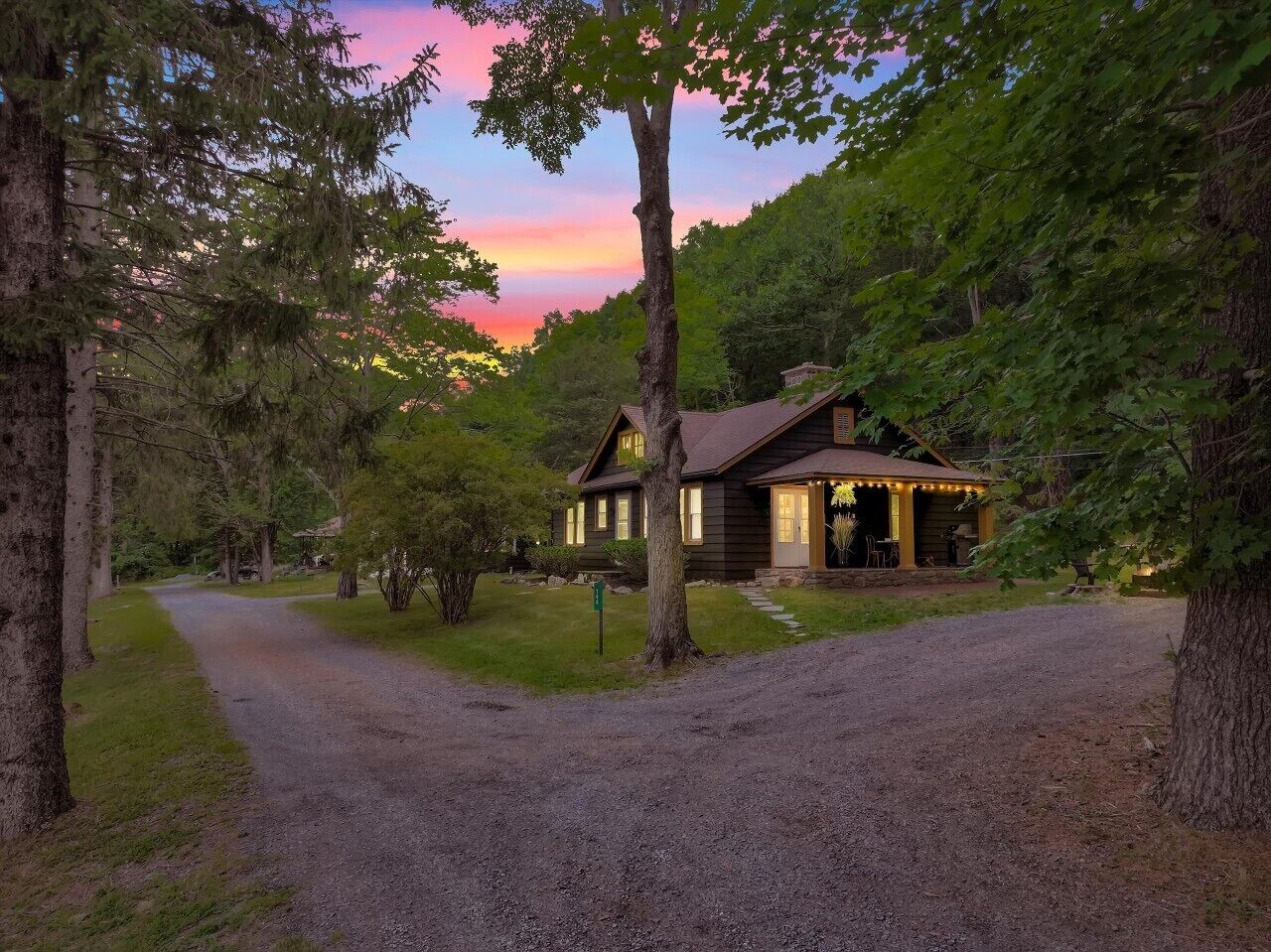 A rustic cabin with a gravel driveway, surrounded by trees and a sunset in the background.