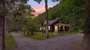 A rustic cabin with a gravel driveway, surrounded by trees and a sunset in the background.