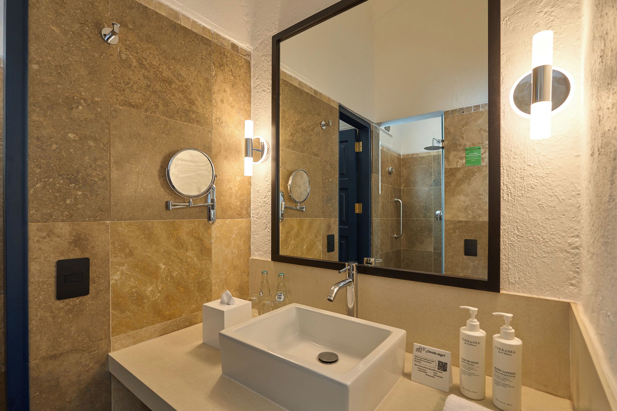 Un baño con un lavamanos blanco, dos espejos, un cabezal de ducha de mano y artículos de aseo.