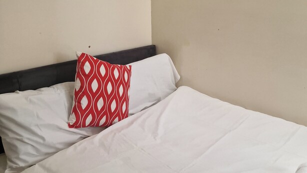 A neatly made bed with white linens and a single red patterned pillow, a rolled white towel, and a plain wall. - A Room Double bed 2 People With Hob and Fridge (London)