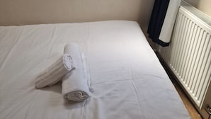 A single bed with white sheets and a rolled-up towel, a white radiator, and a window with a dark curtain. - A Room Double bed 2 People With Hob and Fridge (London)