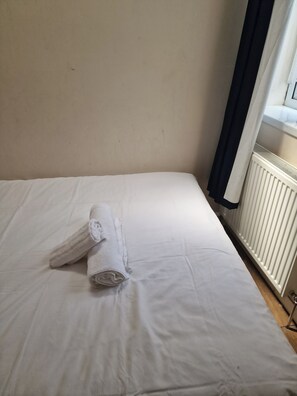 Ein Einzelbett mit weißen Laken und einem zusammengerollten Handtuch, eine weiße Heizung sowie ein Fenster mit dunklem Vorhang.