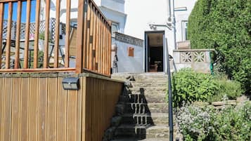 A wooden staircase leading to a white building with a balcony, stone steps, and a garden with flowering plants.
