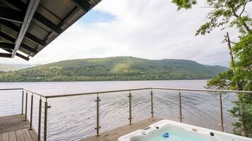 Um deck de madeira com banheira de hidromassagem e vista para um lago e colinas arborizadas.