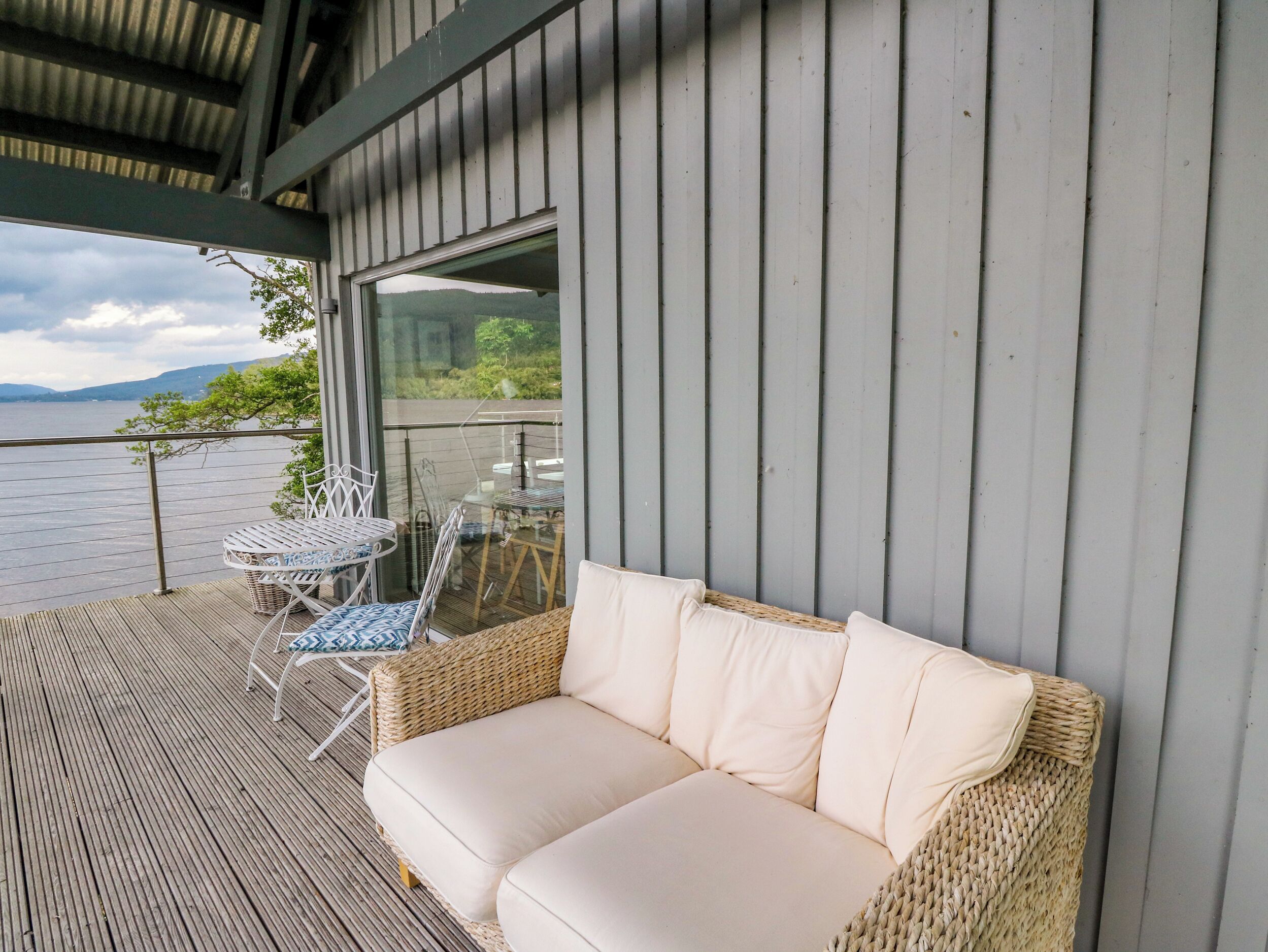 A balcony with a wicker sofa and a table, overlooking a body of water and greenery.
