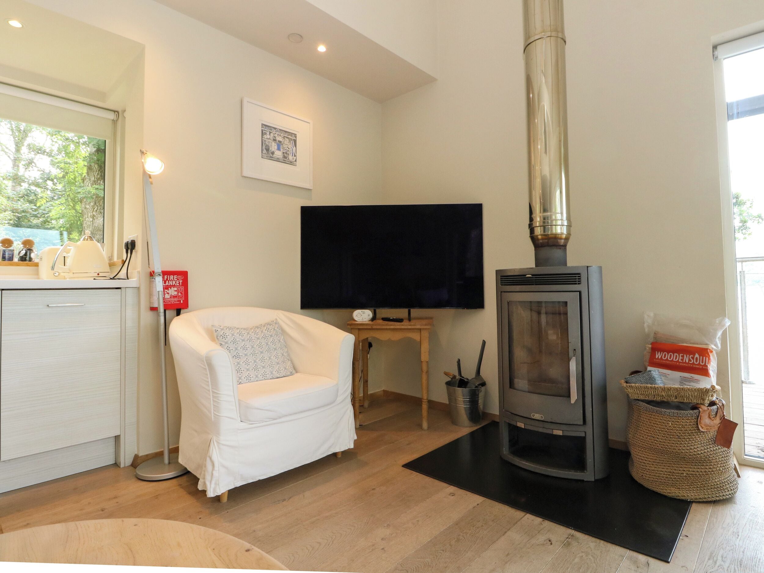 Een gezellige woonkamer met een witte fauteuil, een flatscreen televisie, een houtkachel en een raam met uitzicht op groen.