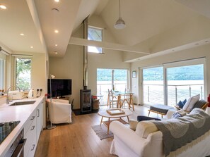 A modern interior with a kitchen, dining area, and living room, featuring a large window with a view of the water.