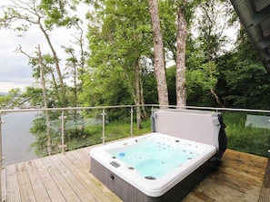 A hot tub on a wooden deck with a glass railing, surrounded by trees and overlooking a body of water.