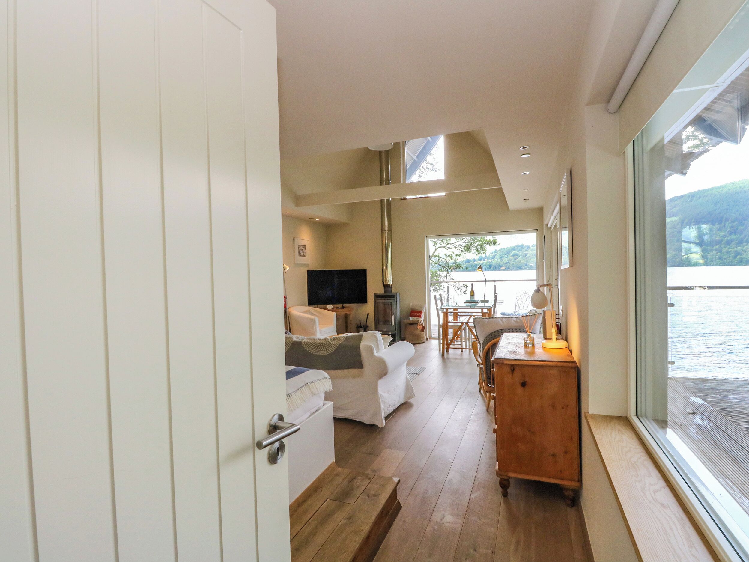 A modern interior with a wooden floor, white furniture, a TV, and a view of a body of water.