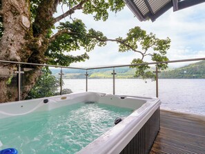 Een hottub met uitzicht op een meer en heuvels, omringd door een reling en een boom.