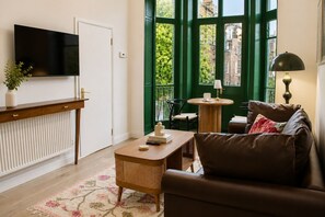 Ein Wohnzimmer mit Fernseher, einer Couch, einem Couchtisch und Blick auf ein grünes Gebäude durch ein Fenster.