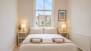 A bedroom with a large bed, two bedside tables, a window with a view of buildings, and a framed picture on the wall. - Notting Hill's Iconic Pink Apartment (London)