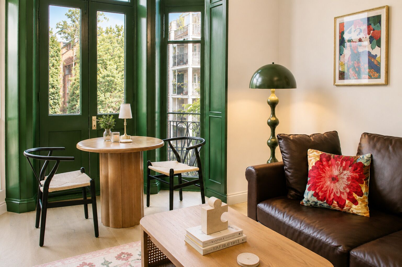 A cozy living room with a green door, a round wooden table, two chairs, a leather sofa with a floral pillow, a floor lamp, and a framed artwork on the wall.
