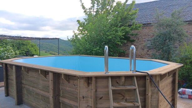 A wooden above-ground swimming pool with two metal ladders, surrounded by greenery and a stone building. - A spacious vacation rental in the countryside with a magnificent view of the Muse Valley. (Saint-Beauzély)
