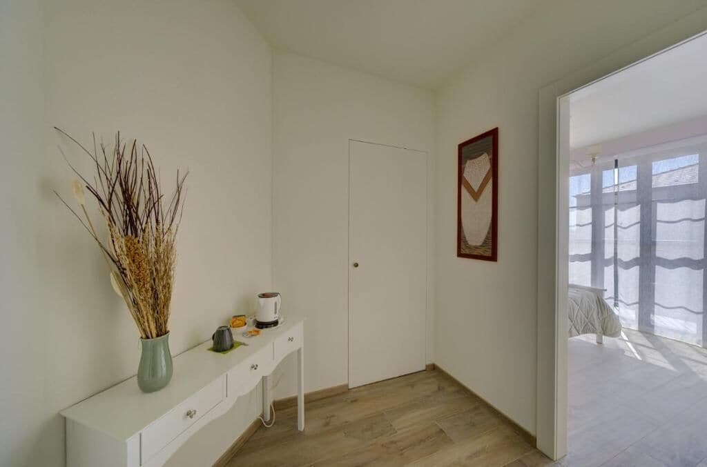 A minimalist room with a white console table, a vase with dried plants, a coffee maker, and a door leading to a balcony with a view.