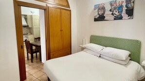 A bedroom with a bed, a green headboard, a wall-mounted painting, a wooden wardrobe, and a dining area visible through an open door. - PILLYHOUSE VUCCIRIA: a charming apartment at the entrance to the Vucciria Market (Palermo)