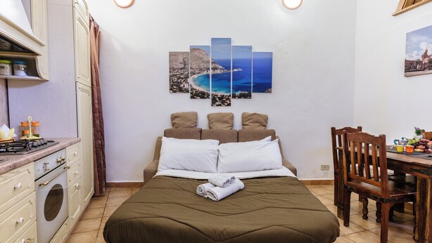 A bedroom with a bed, a dining table, a kitchen area, and a wall-mounted artwork. - PILLYHOUSE VUCCIRIA: a charming apartment at the entrance to the Vucciria Market (Palermo)