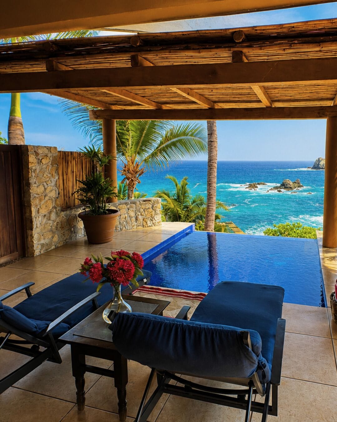 An infinity pool with a view of the ocean, a stone wall, and a wooden pergola.