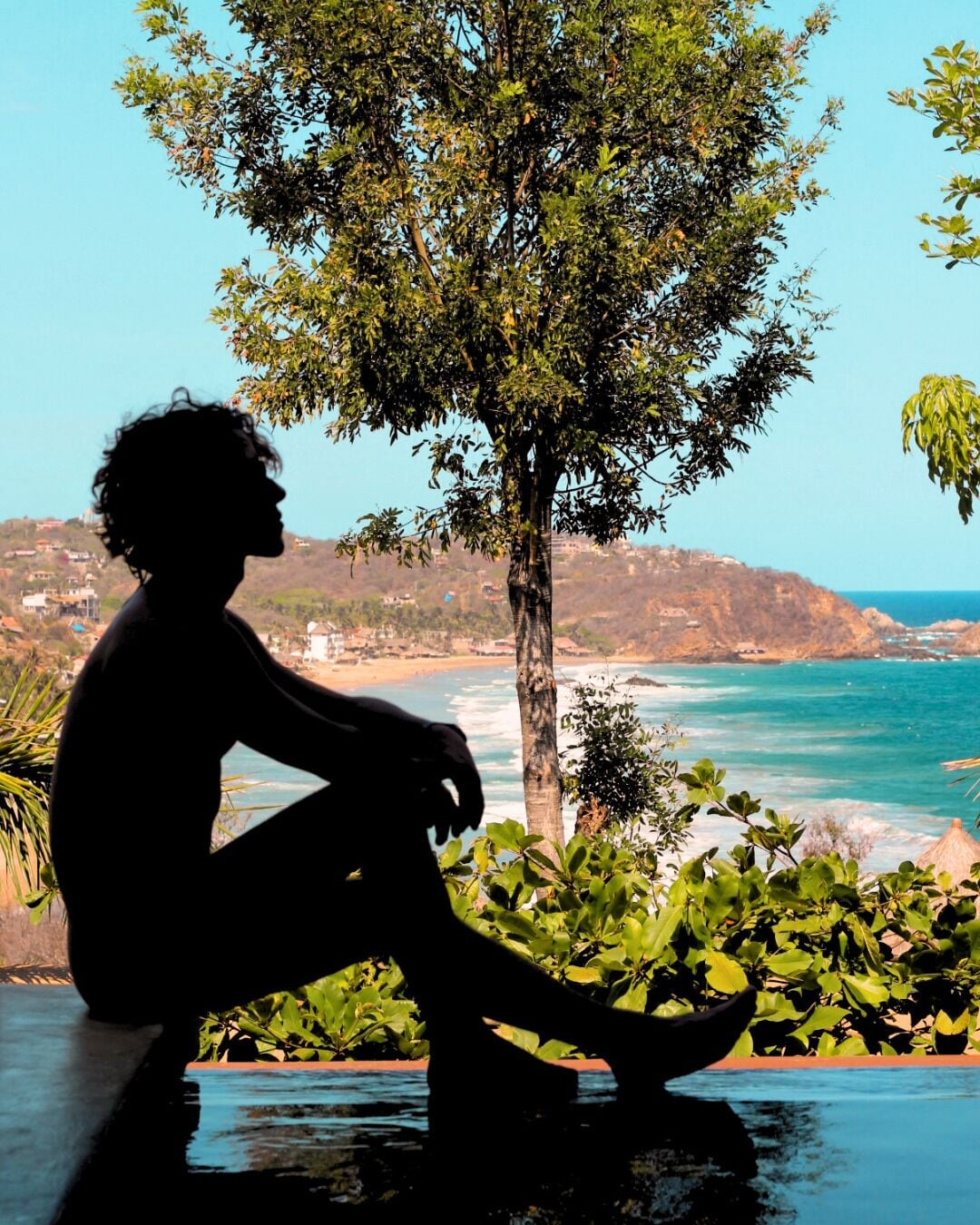 Una persona sentada junto a una piscina con vistas a una playa y al océano.