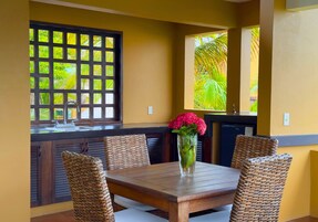 Un comedor con una mesa de madera y sillas de mimbre, un florero con flores y vistas al verdor a través de una ventana.