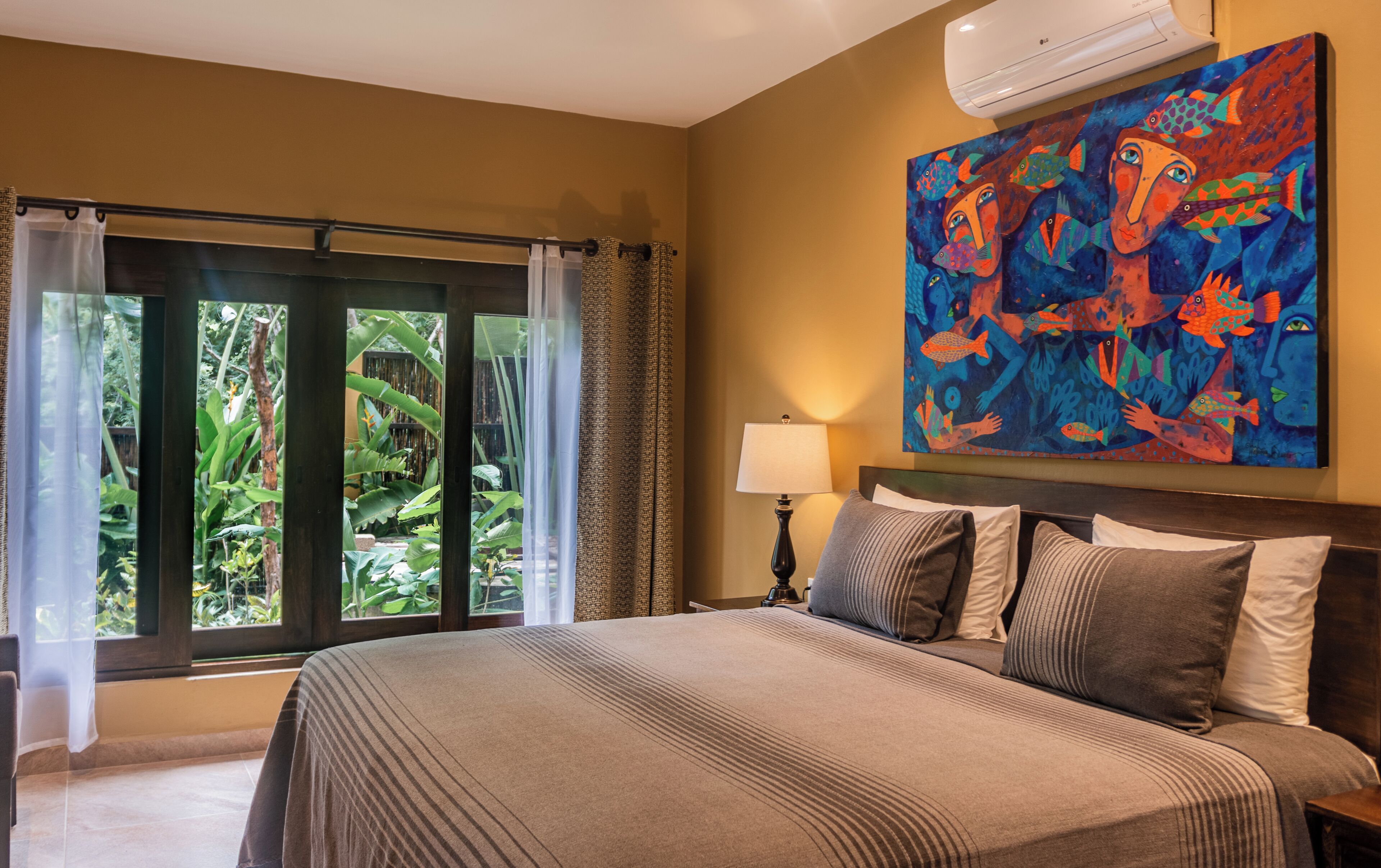 A hotel room with a large bed, a painting on the wall, a window with curtains, and an air conditioning unit.