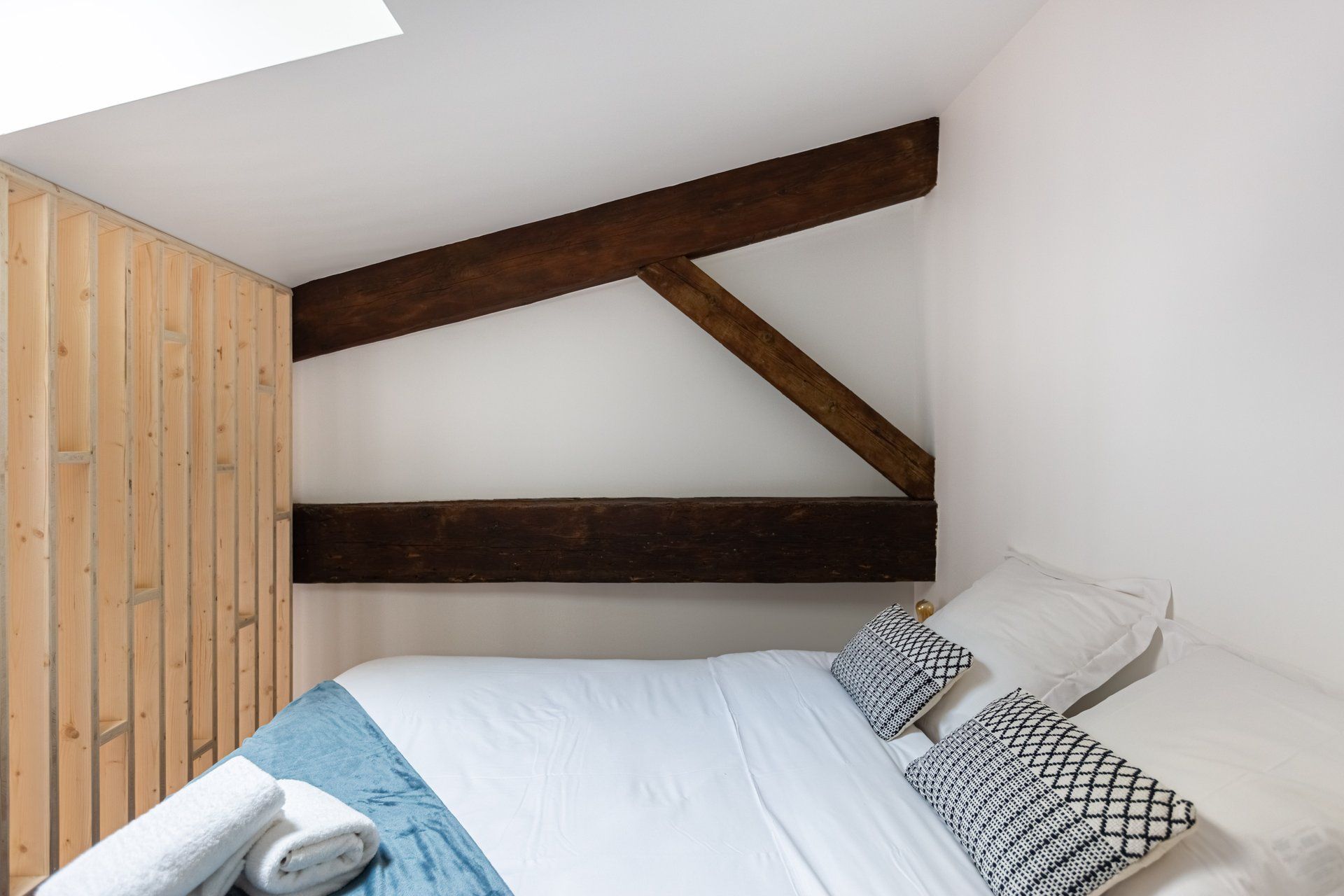 A small, cozy room with a sloped ceiling, a wooden wall, and a bed with white bedding and patterned pillows.