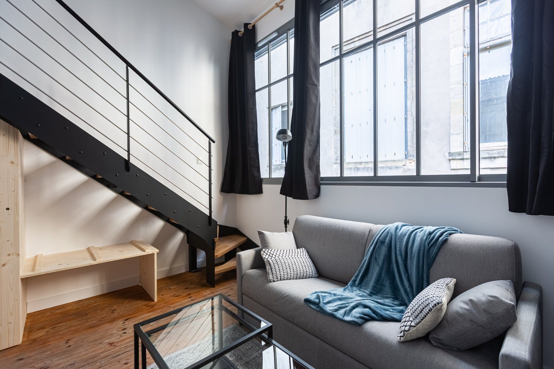 Een moderne woonkamer met een grijze bank, een glazen salontafel, een houten bankje en een zwarte trap.