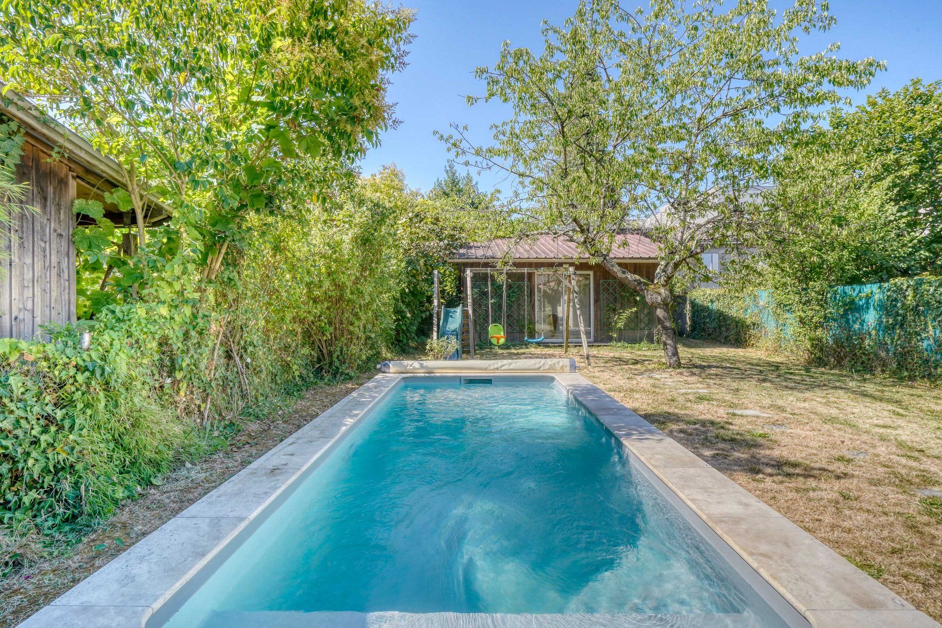 Piscina rectangular con borde de hormigón, rodeada de césped y árboles.