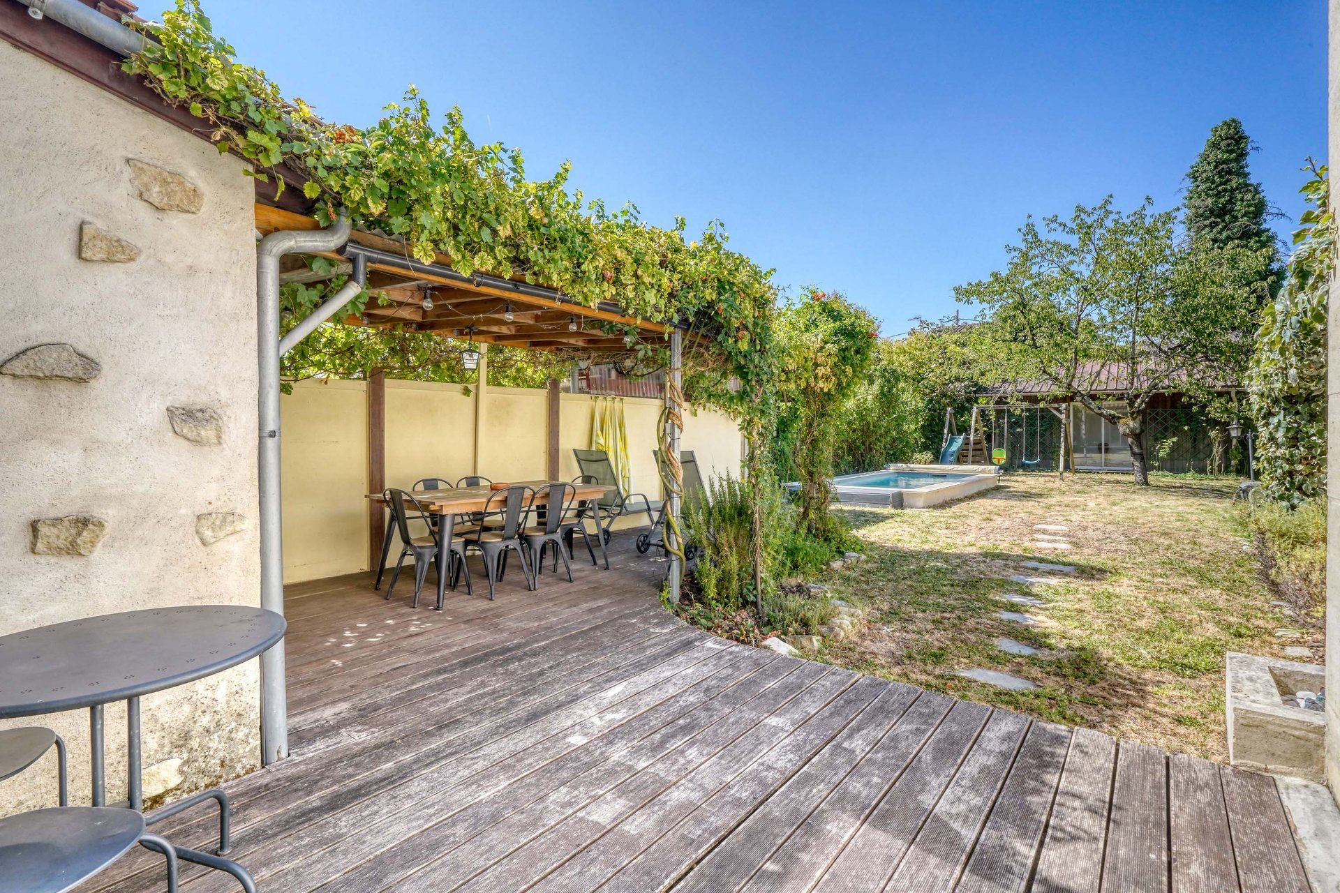 Une terrasse extérieure avec une plateforme en bois, une table et des chaises, et une pergola recouverte de plantes grimpantes.