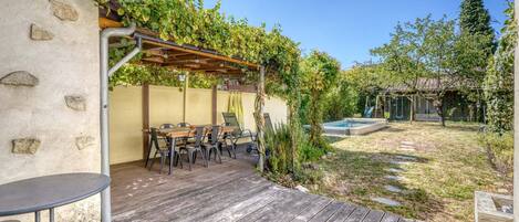 Un patio exterior con terraza de madera, una mesa con sillas y una pérgola cubierta de enredaderas.