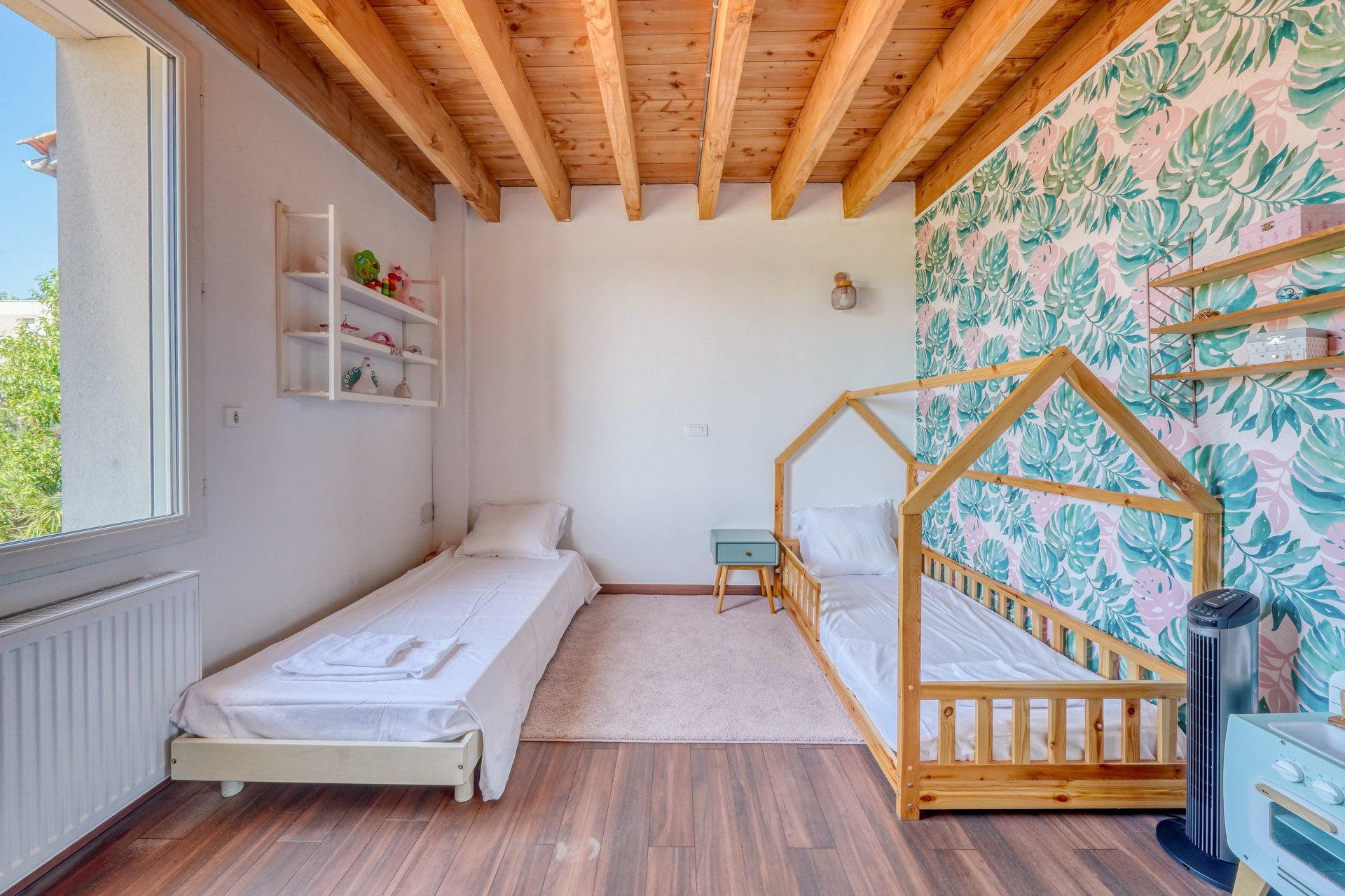 A bedroom with two single beds, a wooden ceiling, a radiator, a shelf with toys, and a wall with a leafy pattern.