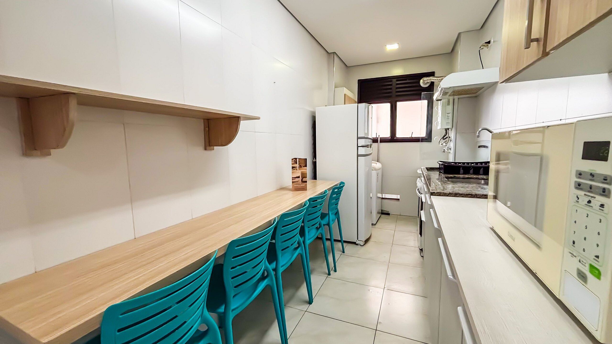 Una cocina con una mesa de madera, sillas color turquesa, armarios blancos, un refrigerador y un microondas.