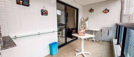 Un petit balcon avec une table, une poubelle et une étagère en verre adossée à un mur recouvert de carreaux blancs.