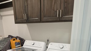 A laundry area with two white front-loading washing machines, wooden cabinets above, and a yellow detergent bottle on the left. - Ash & Alder Townhouse (Idaho Falls)