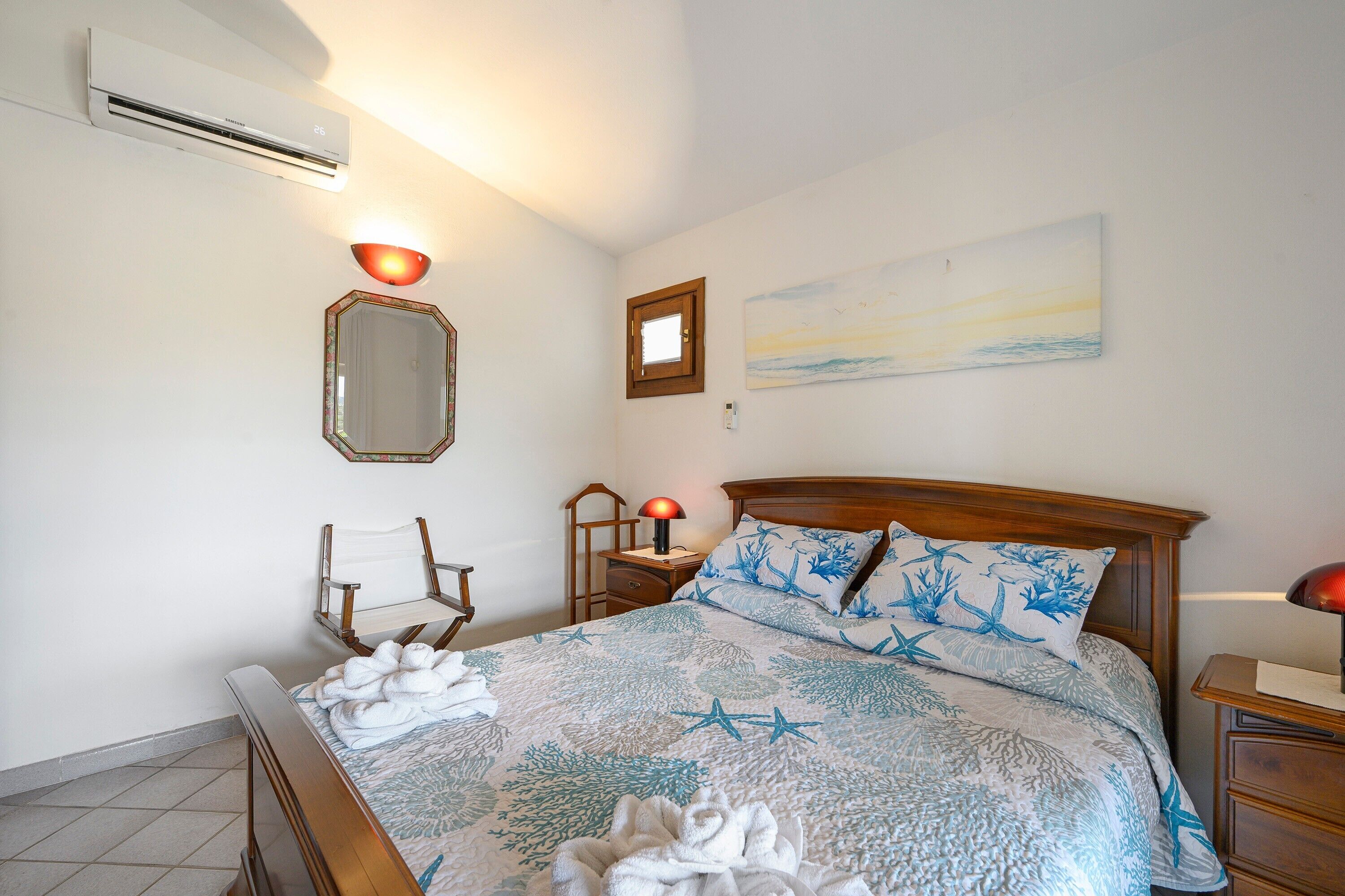 Une chambre à coucher comprenant un lit, une table de chevet, une chaise, un miroir, une lampe fixée au mur et un tableau.