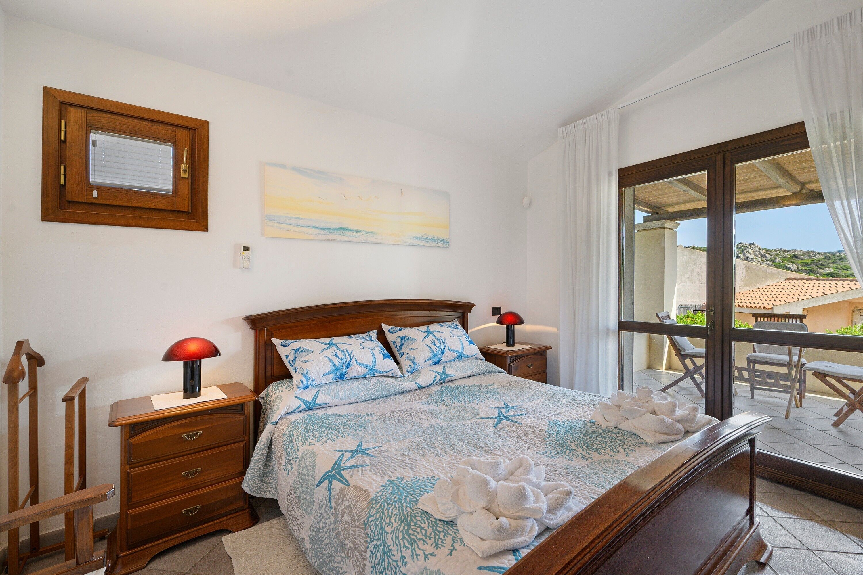 Une chambre à coucher avec un lit, des tables de chevet en bois, un tableau et une vue sur l’extérieur.