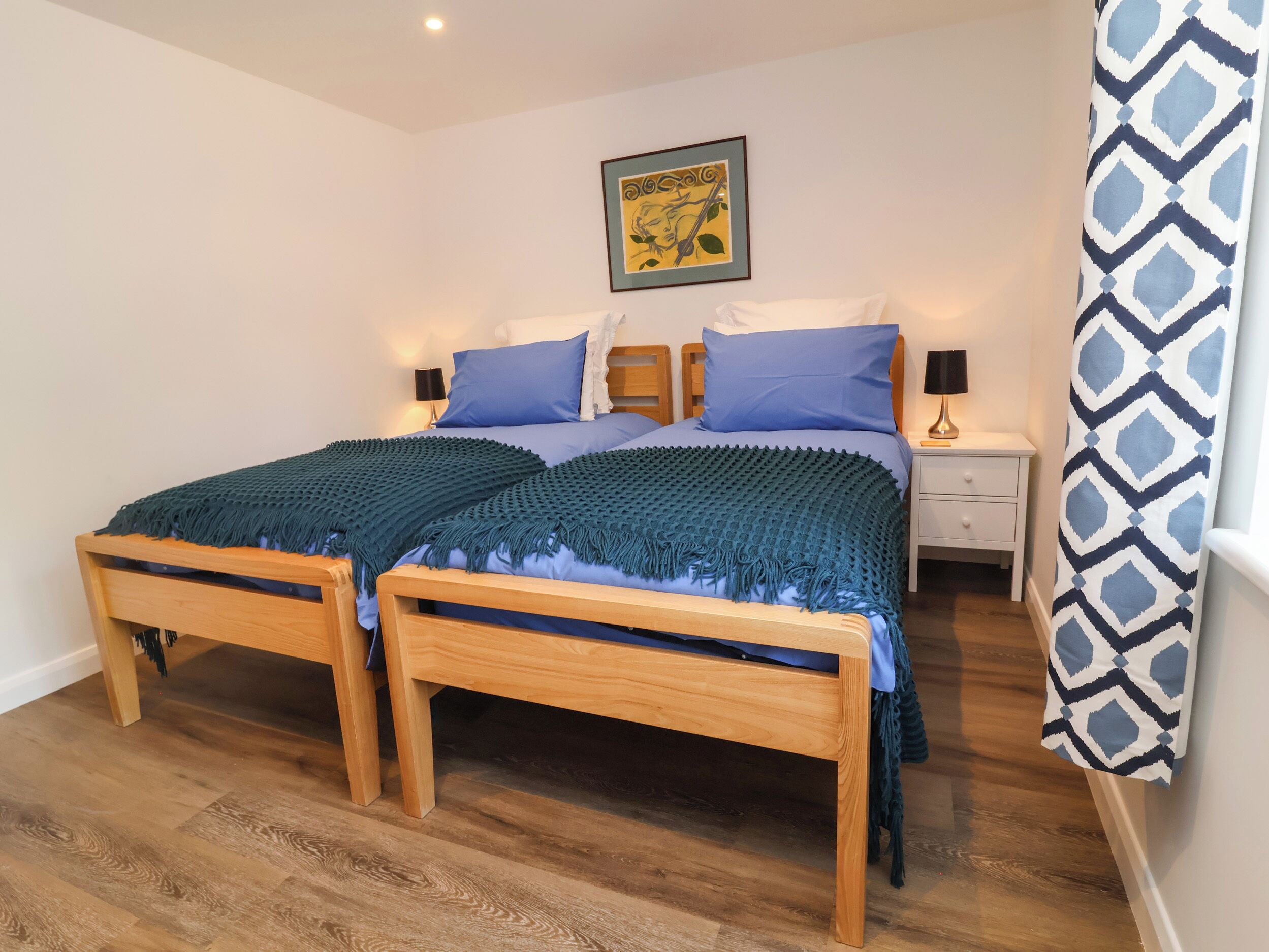 A double bed with a blue and green blanket, two bedside tables with lamps, a framed picture on the wall, and a patterned curtain.
