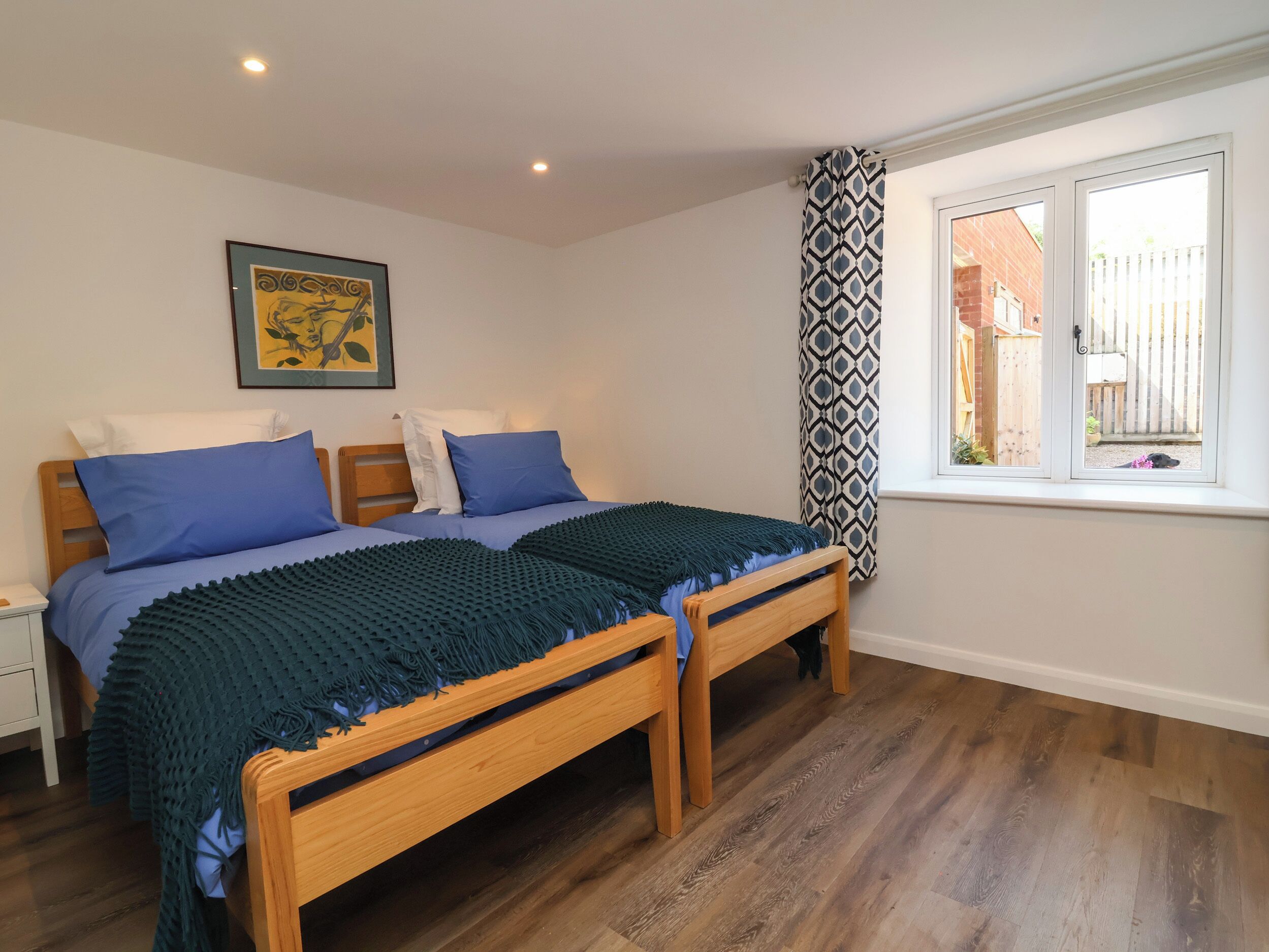 Un dormitorio con dos camas de madera, una ventana con cortinas estampadas y un cuadro en la pared.