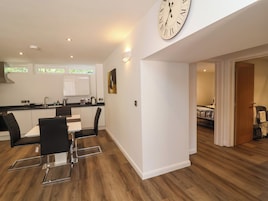 A modern kitchen with a dining area, a large wall clock, and a view into a bedroom.