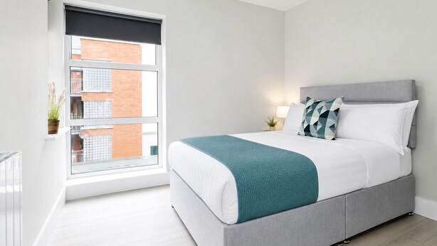 Ein modernes Schlafzimmer mit Bett, Nachttisch, Lampe und Blick auf ein Gebäude durch ein Fenster.