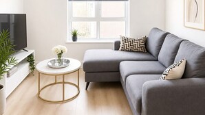 Ein modernes Wohnzimmer mit grauem Sofa, einem runden Couchtisch, einem Fernseher und einem Fenster mit Jalousien.
