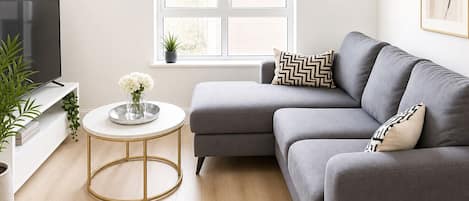 A modern living room with a grey sofa, a round coffee table, a TV, and a window with blinds.