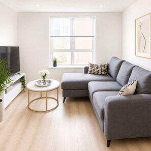 A modern living room with a grey sofa, a round coffee table, a TV, and a window with blinds. - Modern Central Dublin Apartment Near Bolton Square (Dublin)