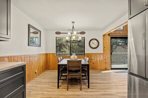 Un coin repas comprenant une table en bois, des chaises, un lustre, une horloge et une fenêtre avec des stores.