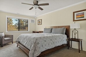 A bedroom with a bed, a chair, a nightstand, a lamp, a ceiling fan, and a window with a view of trees. - Quiet Mountain Cabin Escape (Lake Arrowhead)